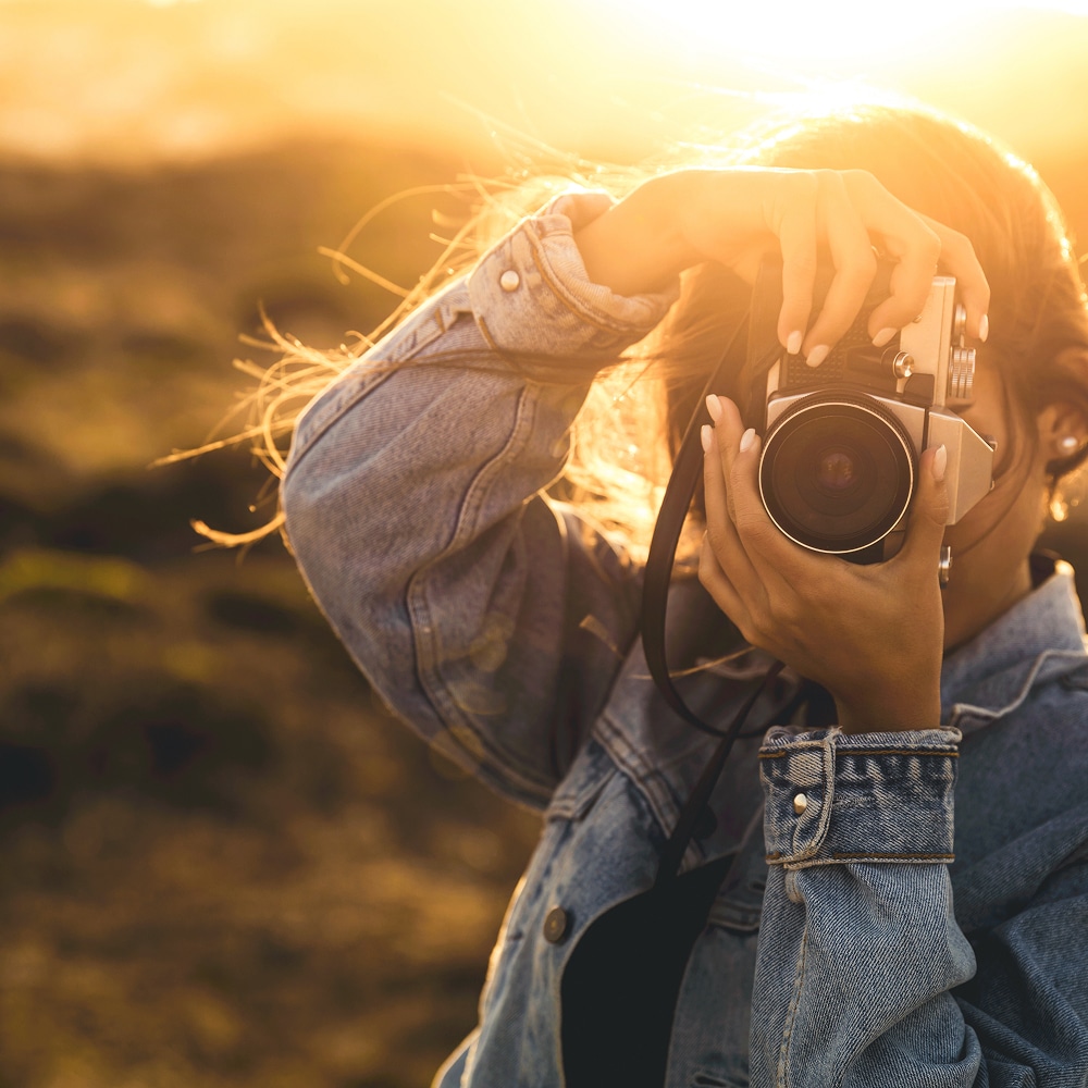 Femme en veste en jean utilisant un appareil photo vintage sous la lumière dorée d'un coucher de soleil.