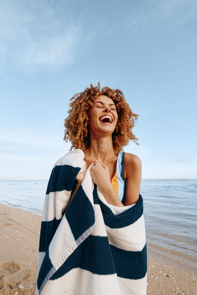 Femme rieuse aux cheveux bouclés sur une plage, enveloppée dans une serviette rayée bleue et blanche sous un ciel dégagé.