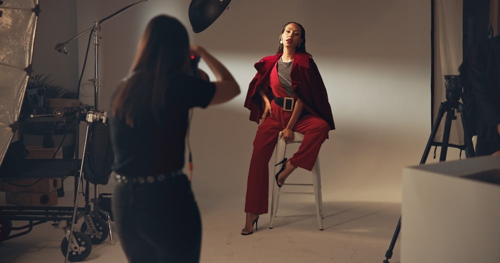 Photographe capturant un mannequin en tenue rouge sur un tabouret lors d'un shooting de mode en studio professionnel.